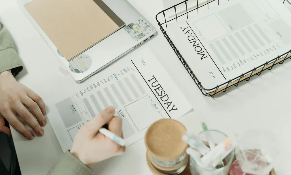 Person planning a weekly cleaning schedule by writing in a Tuesday planner on a desk with laptop and Monday schedule